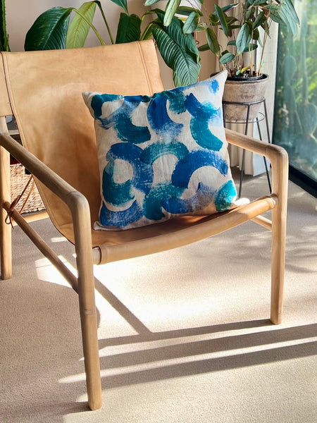 A square throw pillow with a blue and white abstract brushstroke pattern, resting on a light tan leather and wood lounge chair in a sunlit room with potted plants.