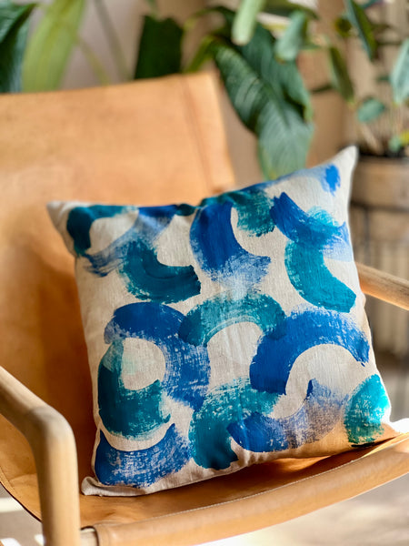 A square throw pillow with an abstract blue and teal brushstroke pattern on a cream linen background, styled on a light wood chair with a blurred indoor plant background.