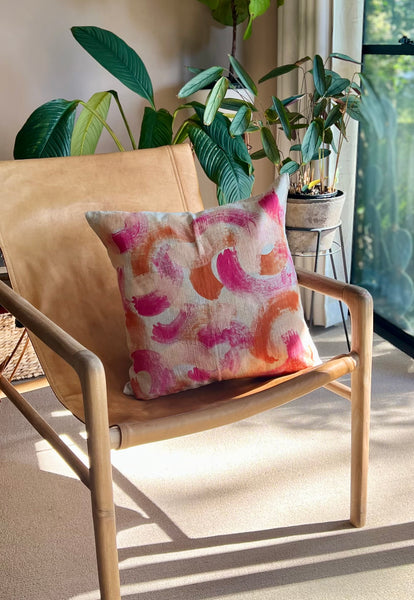 A square throw pillow with a pink, orange, and white abstract brushstroke pattern, resting on a tan leather sling armchair in a sunlit room with potted plants.