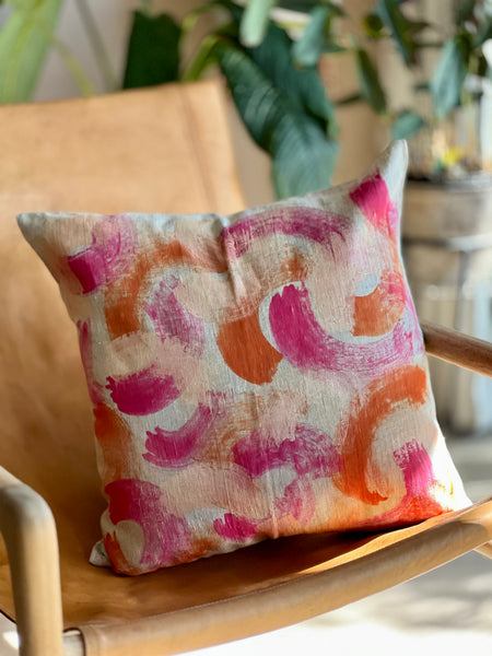 A square throw pillow with an abstract watercolor brushstroke pattern in pink, orange, and white, resting on a light brown wooden chair with a blurred background of indoor plants.