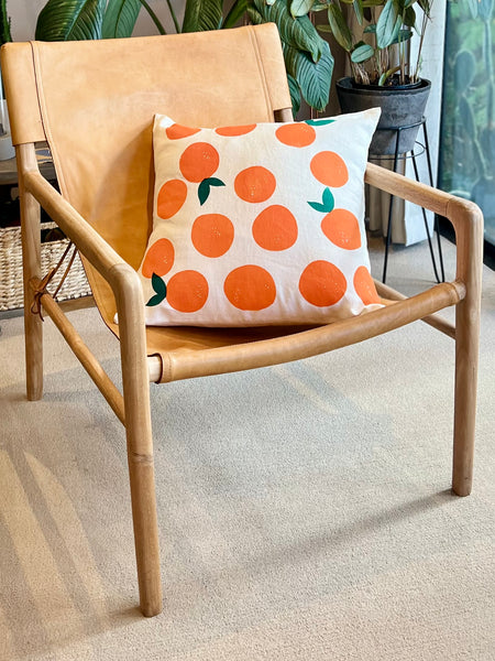 A light natural wood armchair with a tan leather sling seat and back, styled with a square throw pillow featuring a white background and orange citrus fruit pattern with small green leaves.