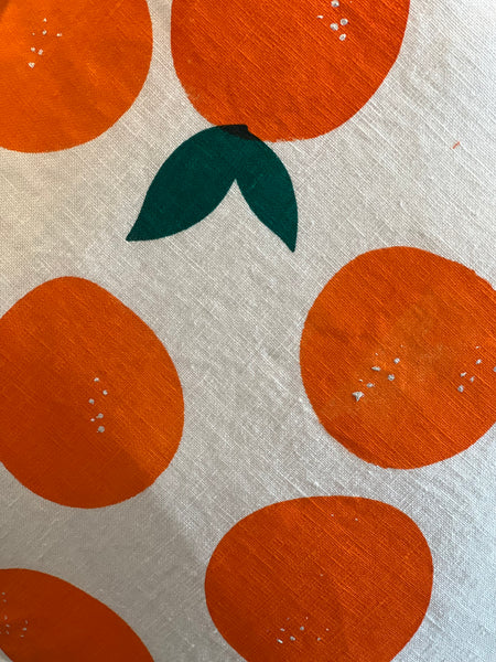 A close-up of a fabric surface with a repeating orange citrus fruit print on a light beige background, featuring dark green leaves.