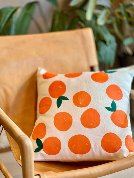 A square throw pillow with a pattern of orange oranges and green leaves on a cream background, resting on a light wood chair with a blurred indoor plant in the background.