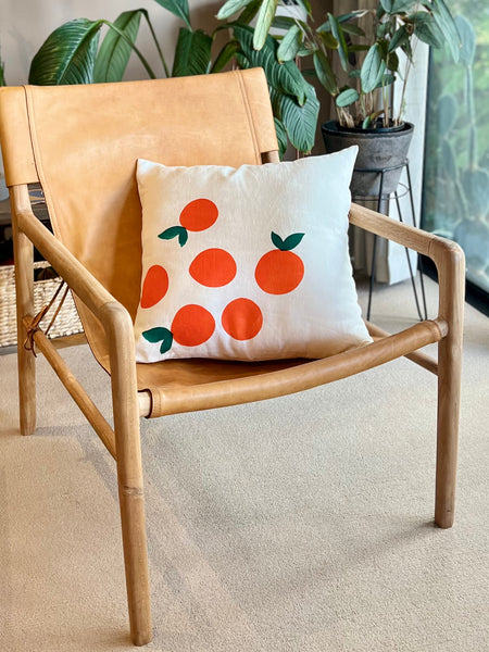A square throw pillow with a bold orange and green citrus fruit pattern on a cream background, styled on a light tan leather and wood armchair in a bright room with potted plants.