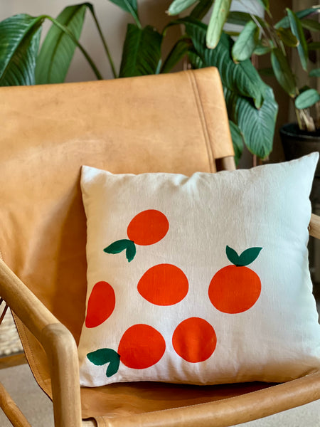 A square throw pillow with a pattern of orange fruits and green leaves, resting on a tan leather chair with green houseplants in the background.